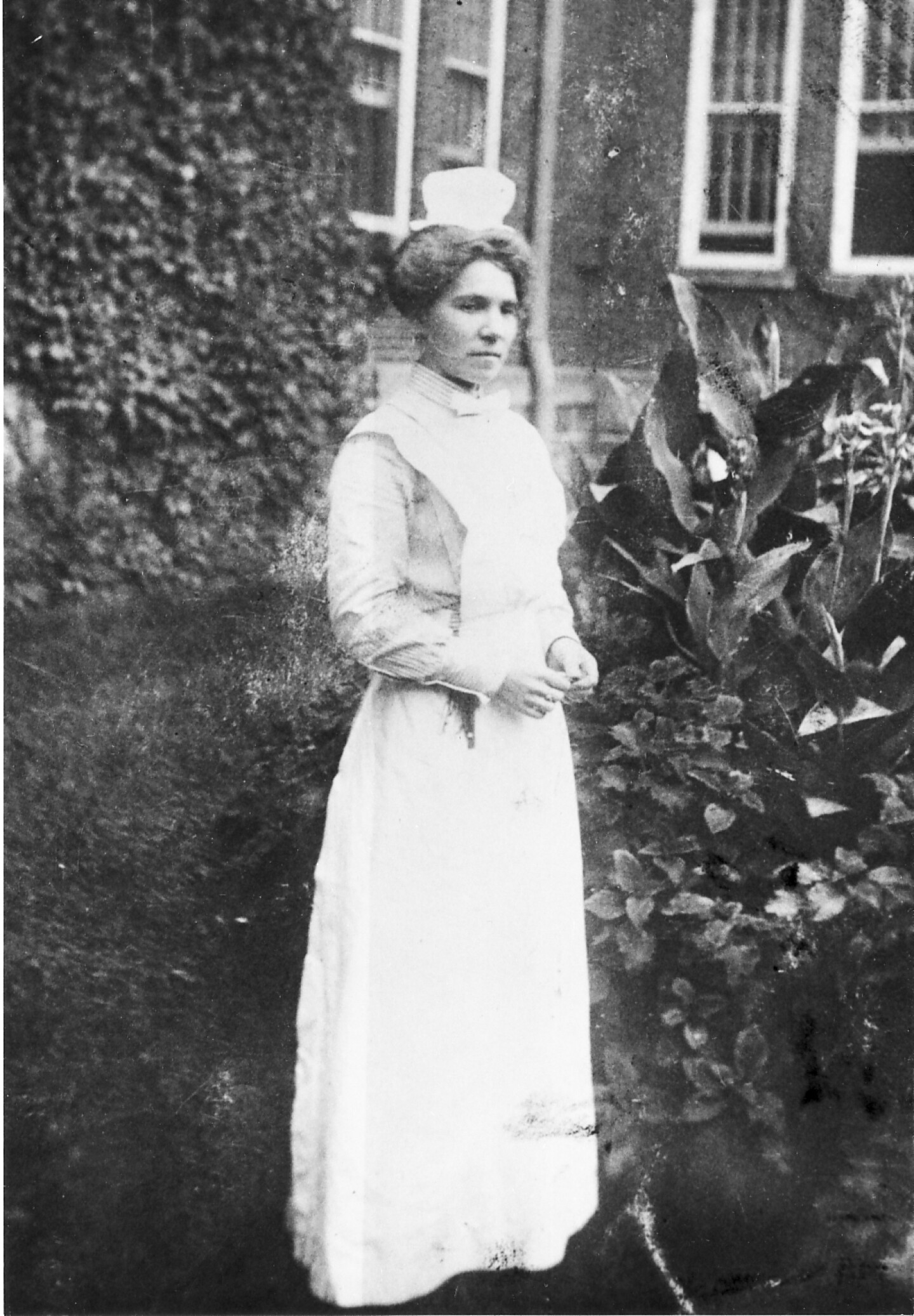 An undated photo of Rockhaven Sanitarium founder Agnes Richards standing in the Sanitarium's garden. She is wearing a white dress with a bib over a blouse. She has what appears to be a type of nurse's cap on her head.