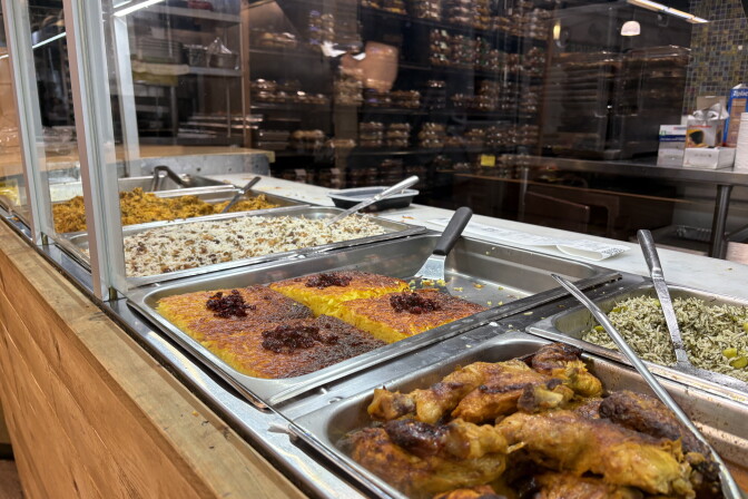 An array of metal serving containers has different foods in each, with long serving spoons. They're in a wooden section in a supermarket.