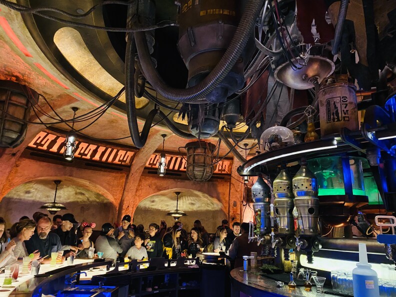 Many people stand and sit near a circular bar inside a large bar. The walls are brown and curved. Mechanical equipment and wires hang from the ceiling.