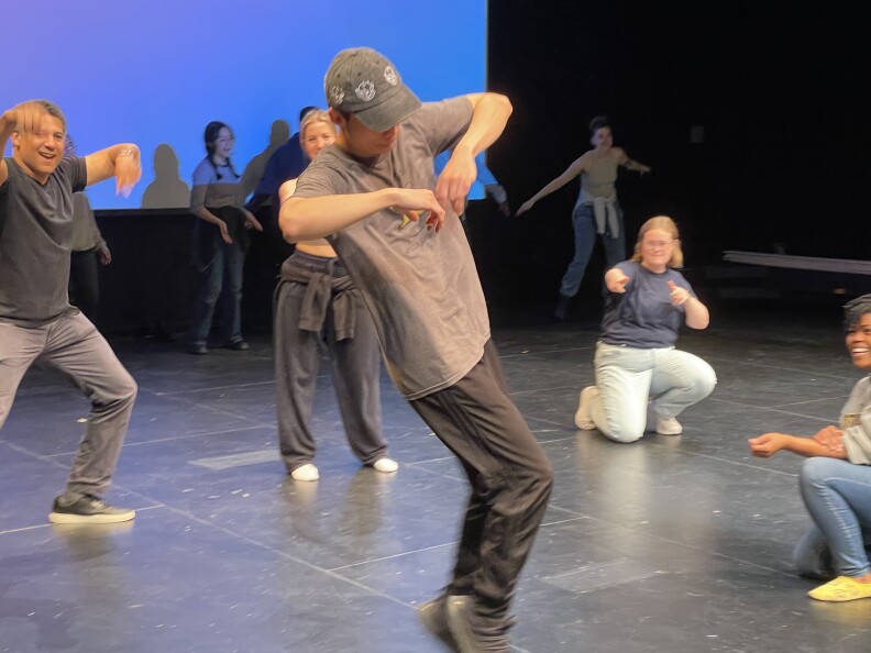 A man with light skin and a dark cap dances in a room with others. He is wearing black pants and a gray T-shirt. He is in mid-dance with his arms up an bent inward at the elbows. His head is down and his legs are mid-hop to his left.