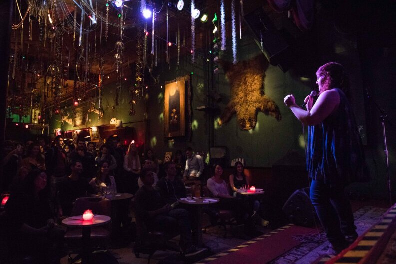 A woman performing stand up in a dimly lit bar, facing a rather packed crowd. Some people in the crowd are smiling and laughing. The woman has the spot light on her while everyone else in the crowd is in the dim lighting and paying attention to her.