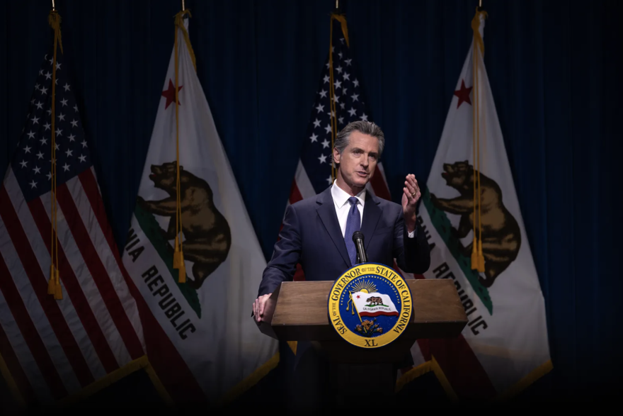 A man with light skin tone dressed in a dark blue suit is at a podium and speaking. Behind him are flags for the United States as well as California.