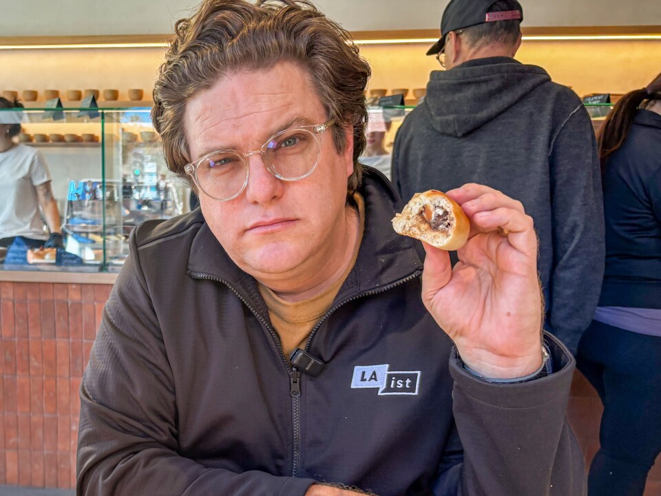 A man with a light skin tone and wavy brown hair holds up a piece of bagel inside Miopane bakery.