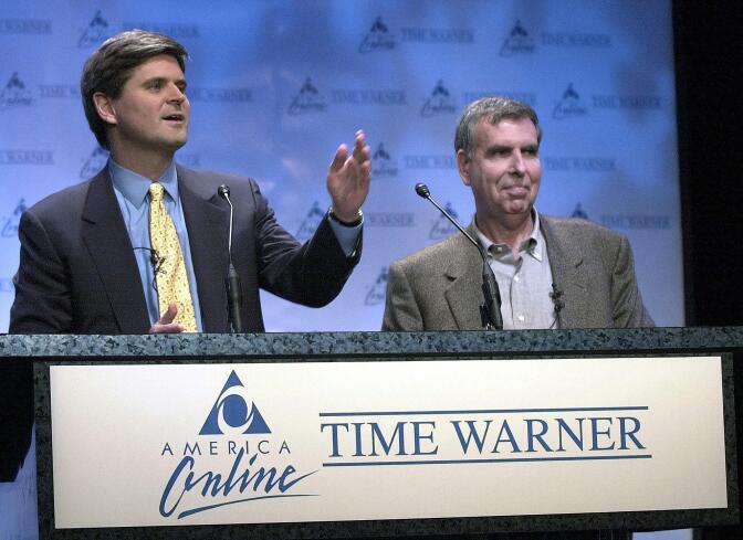 America Online Chairman Steve Case, a man with light skin tone, wearing an indigo suit, speaks behind a podium as he stands next to Time Warner Chairman Gerald Levin, a man with light skin tone, wearing a gray charcoal suit. The podium has signage that of past America Online and Time Warner logos.