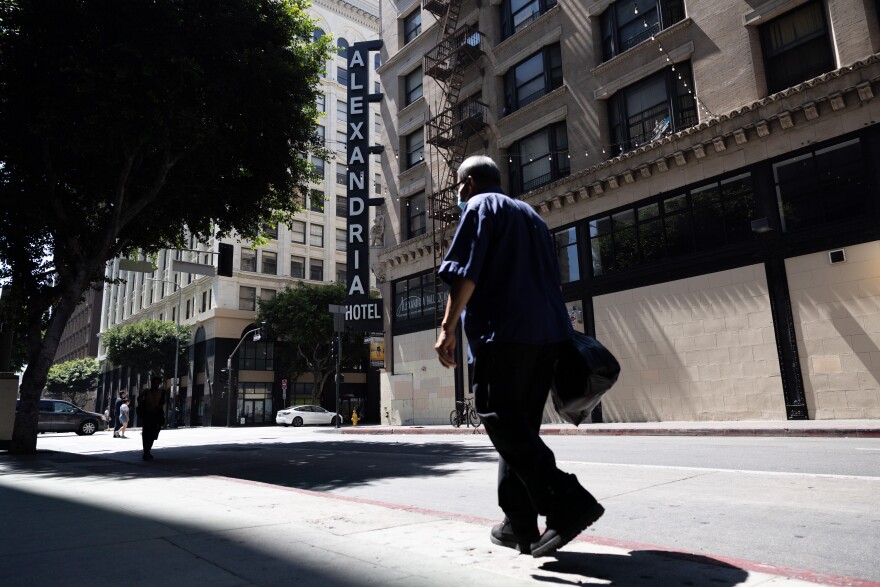 A man walks along a sidewalk. In front of him is a blade-style sign on the side of a building that reads "Alexandria Hotel."