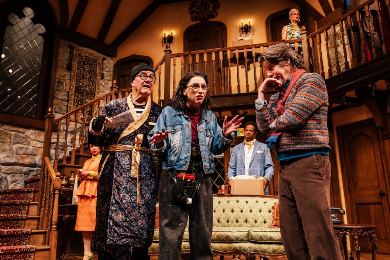Six actors, men and women of varying races and ages, stand on stage in a production of "Noises Off." The set is the interior of a home with a varnished wooden staircase and stone wall.