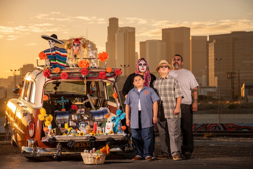 A man, a woman and two children stand next to a vintage station wagon decorated with Dia de los Muertos paraphernalia. In the background is downtown L.A. The sun is setting.