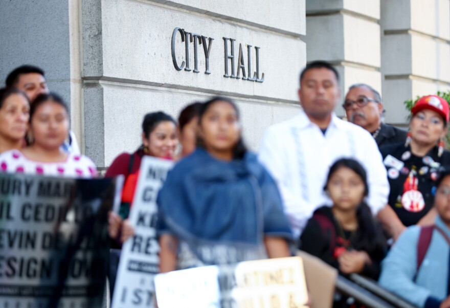 In the foreground, a group of people with medium skin tones hold protest signs calling for Nury Martinez, Gill Cedillo and Kevin de León to resign. In the background is a building that says City Hall.
