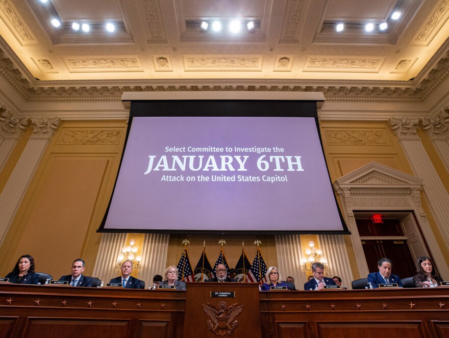 9 people site behind a long wooden desk, 4 American flags stand behind the middle three. Above them is a screen depending from the ceiling with a blue background and the words "Select Committee to Investigate the JANUARY 6TH Attack on the United States Capitol."