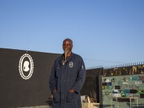 A Black man with a long white goatee wearing a long navy painter's coat, smiles at the camera with his hands in his front coat pockets. Behind him is a black wall with a white, curio-style portrait of him painted on it, and directly behind him is a cinder block wall with each block painted differently. Above him is a clear blue sky.