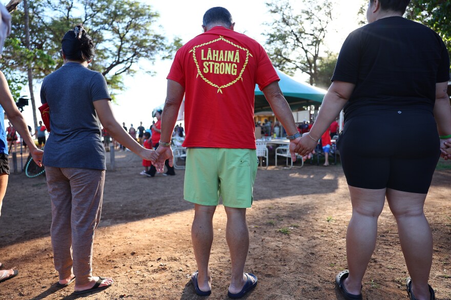 Three people stand, holding hands on a patch of ground outdoors. They are surrounded by trees, people and a tent with table and chairs. The man in the center of the image is wearing green shorts and a red t-shirt that reads "Lahaina Strong" in yellow lettering. He is tan with short, shaved black hair.