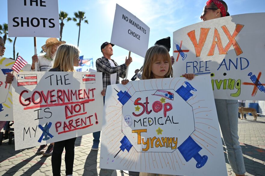 A group of people hold protest signs outside. There is a mix of older adults and children with signs that say messages like, "Stop medical tyranny" and "The government is not my parent!"