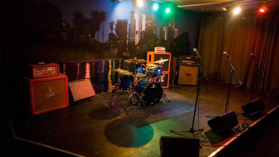 A small black stage with a curtain on one side, a cityscape backdrop on the wall. The stage has drums, amps, speaker monitors, and three microphones on stands.