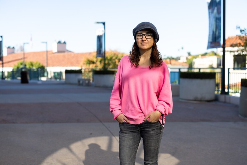 A woman stands outside in the sun wearing dark jeans, a bright pink sweater and a hat.
