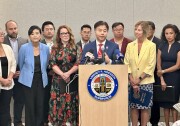 An Asian American man in his 50s speaks at a podium, flanked by ten men and women on both sides of him.