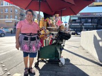 A Brown woman with short curly hair wearing a pink and black apron stand in front of a cart with an umbrella. On the cart, there are lollipops, an assortment of candies and chopped mangoes in plastic containers. 