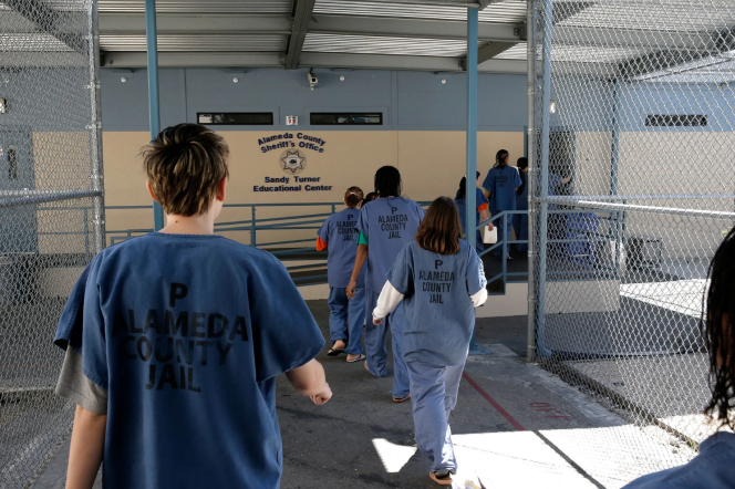 People, wearing blue jumpsuits with text on the back that reads "P Alameda County Jail," walk away from the camera down a chain-link fence gated corridor into a building with signage on it that reads "Alameda County Sheriff's Office. Sandy Turner Educational Center."