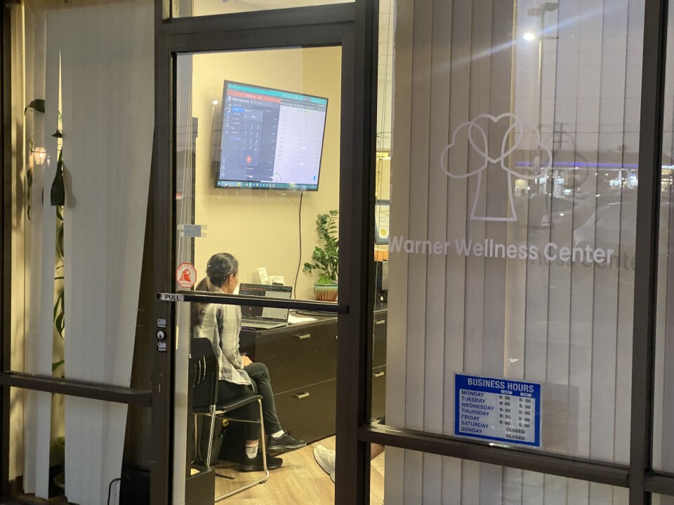 A person sits in an office with facing a TV screen showing a computer screen, with a logo by the door that says Warner Wellness Center