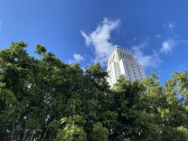 the top of city hall can be seen against a blue sky and clouds with green leafy trees in the foreground. 