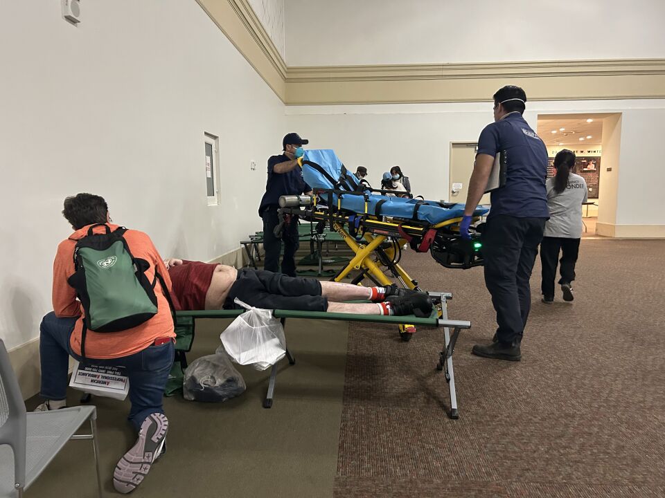 A woman wearing a backback tends to a man laying on a cot, with his bare midriff exposed. Behind them, a pair of EMTs are wheeling a gurney away. 