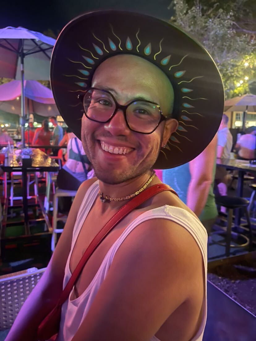 A portrait of Matinez, who's a person with a medium light skin tone wearing a white tank top. They're smiling into the camera wearing dark-rimmed glasses and a round hat.