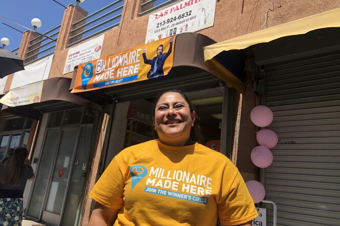 A woman with light brown skin stands in front of a store. She wears a bright yellow t-shirt that says "millionaires made here."