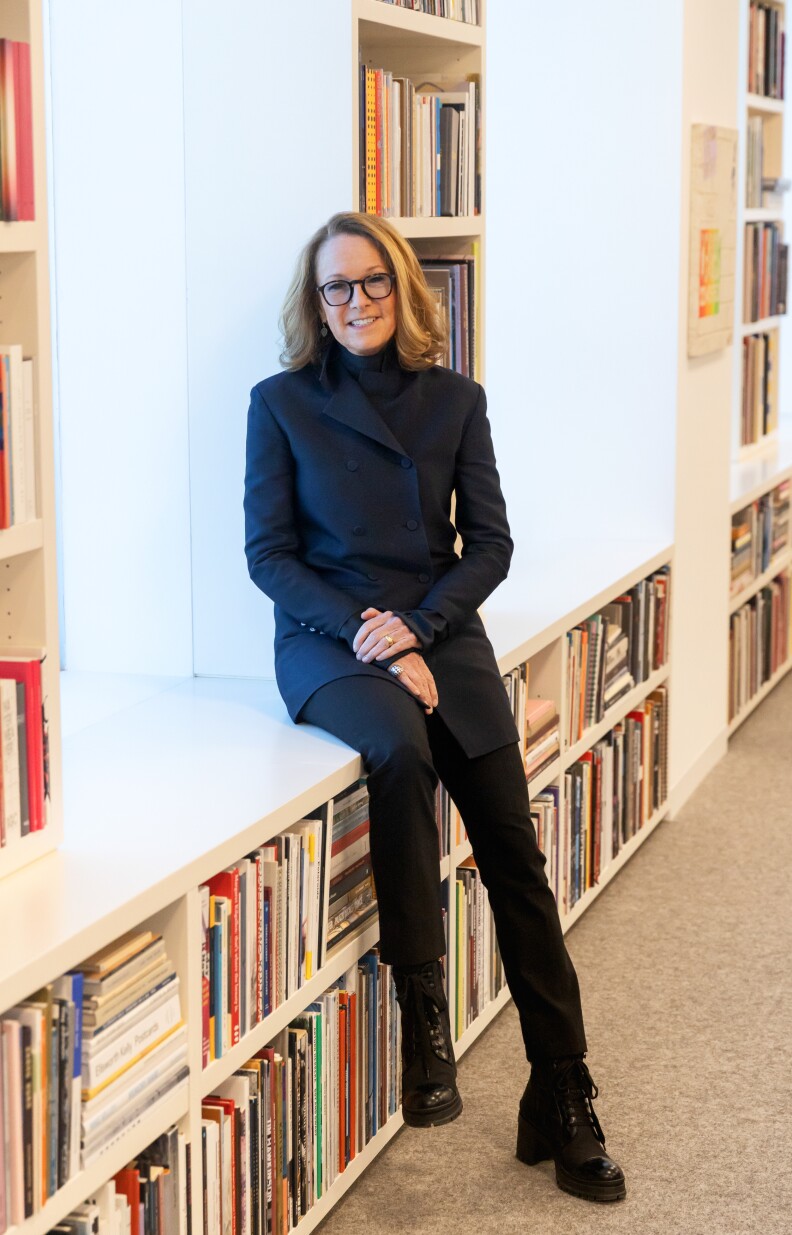 A woman with dirty blond hair wears a black suit and leans on a white shelf of books. Her hands are resting in her lap in front of her.
