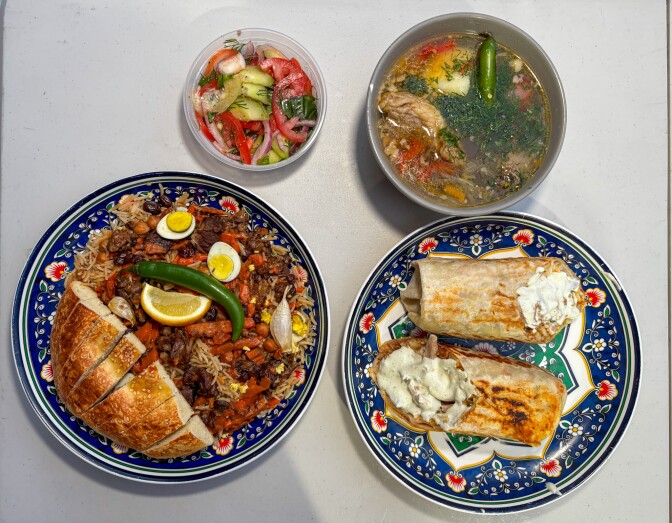 A collection of bowls and plates, all varying in size and contents, is arranged against a pale grey background. At the bottom, there are two plates featuring an intricate blue floral design. The plate on the left holds rice and bread, while the plate on the right contains a large, wrapped item with light brown grill marks and cut ends, topped with white sauce. At the top, there is a small plastic container filled with chopped vegetables resembling a salad, and to its left, a grey bowl holds a liquid mixture of cooked meat, vegetables, and green herbs.