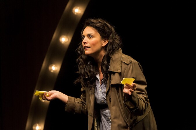 Cecily Strong holds Post-In Notes in both hands while onstage performing in an army green trenchcoat.