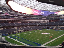 An interior view of the football field at SoFi Stadium in Inglewood. A large, ring-shaped video display board hangs from the roof of the stadium, displaying messages saying the stadium will host Super Bowl 56 on Feb. 13, 2022. Workers on seen preparing the field.