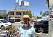 Artist Francisco Palomares looks at the camera. He wears glasses and a large brimmed hat. Behind him is a billboard depicting his oil painting "Fabuloso." The painting depicts a bouquet of orange marigolds in a purple Fabuloso bottle. 
