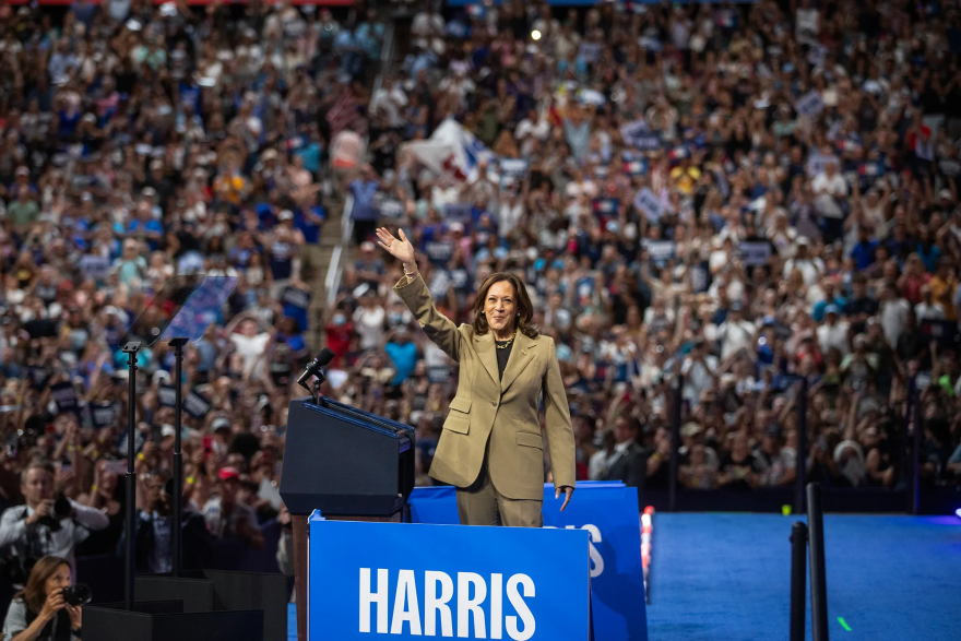 Kamala Harris stands on a stage, waving to a large crowd at a political rally. She is dressed in a tan suit and positioned behind a podium with a blue sign that reads “HARRIS.”