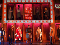 The cast of Assassins are on a stage, several of whom are standing in a two-story cabinet. 