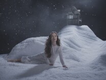 A woman in a white gown lies on a white snow-covered stage scene with a house in the background. 