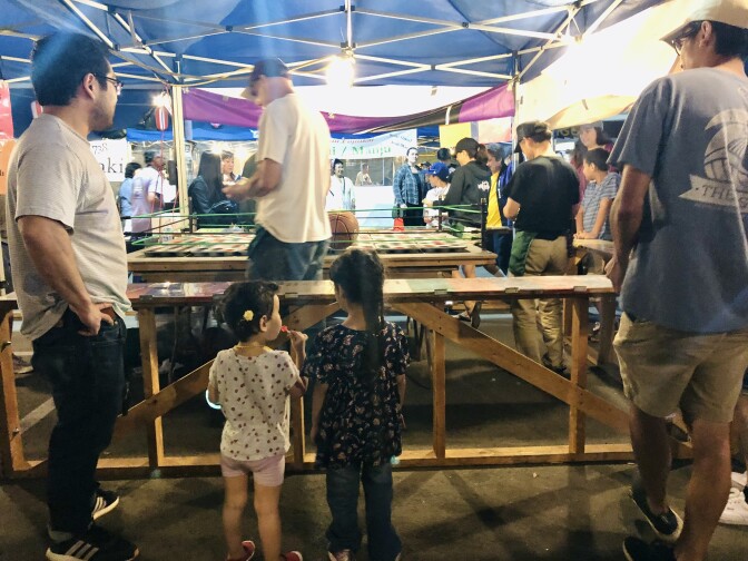 Two young girls look into a game booth. In it, volunteer walks in front of a large wooden table with a basketball and colored cups.