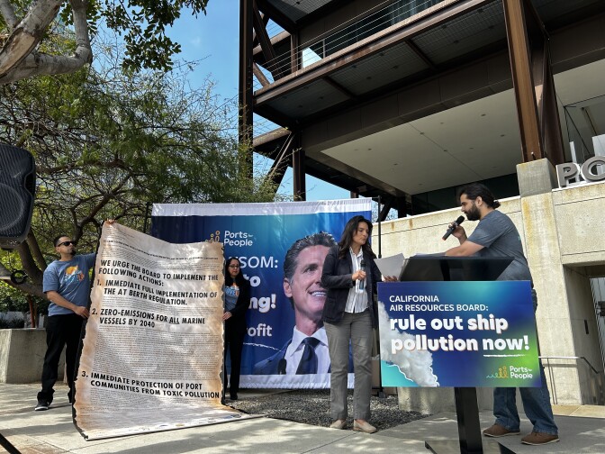 A wide image. To the left, a person with brown skin tone holds an oversized "scroll" with various items on it. To the right are two other people with brown skin tone, one with long hair and another with shorter hair speaking with a microphone at a podium.