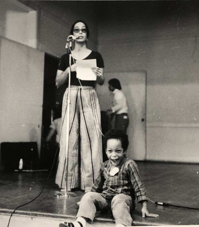 A black and white photo of a Japanese American woman reading at a standing microphone on a stage, while a little boy of Japanese American and Black heritage sits at the edge of the stage.