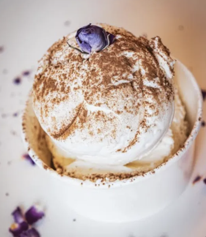A single scoop of white ice cream dusted with cinnamon-colored spice and topped with a small dried purple flower, served in a white paper cup.