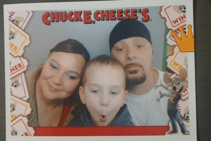 A photograph with a red Chuck E. Cheese's logo across the top and a cartoon mouse in the bottom corner. Two adults and a young child are framed in the photo.