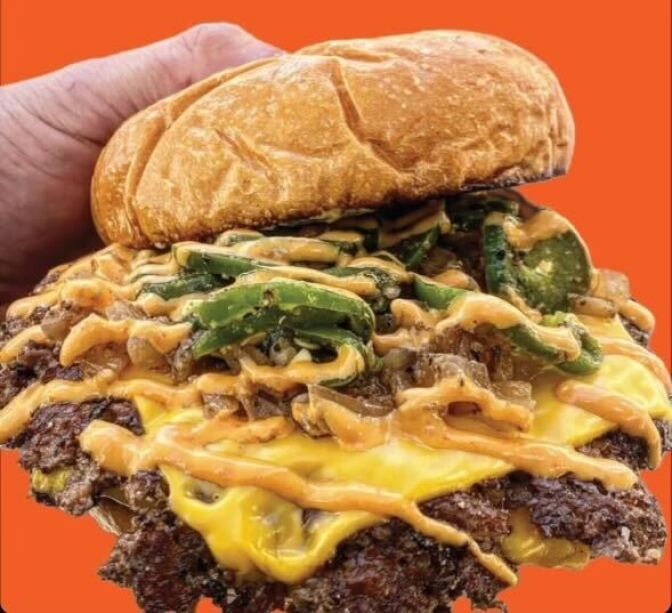 A partially shown light-skinned hand holds a burger stuffed with sliced green jalapeños covered with thin ribbons of orange sauce over a bed of melted American cheese with a  smashed burger patty. 