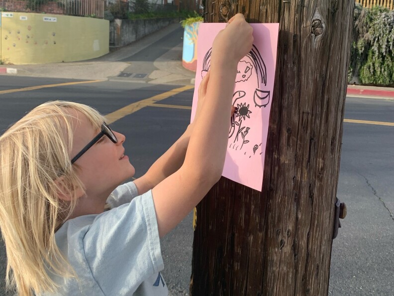 A child with long blonde hair pins a pink handwritten flier to a pole.