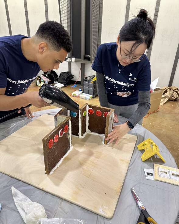 A man and a woman hovering over a gingerbread structure. The man is using a blowdryer to dry frosting holding the pieces together.