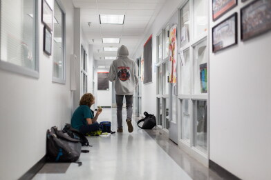 Two students in a bright school hallway: one sits on the floor against the wall eating a snack beside backpacks and a lunch bag, while another in a gray hooded sweatshirt walks away from the camera.