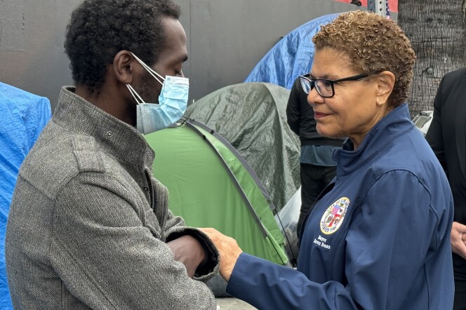 A woman with short hair (right) wearing a blue jacket with the L.A. city seal on it reaches out and holds the arm of a man (left) who's closing his eyes and wearing a mask, in front of tents in the background.