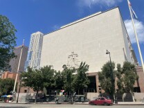 A government building sits on a tree-lined street in downtown Los Angeles, with skyscrapers in the background. Two people pause to speak with someone in a car parked in front of the building. 