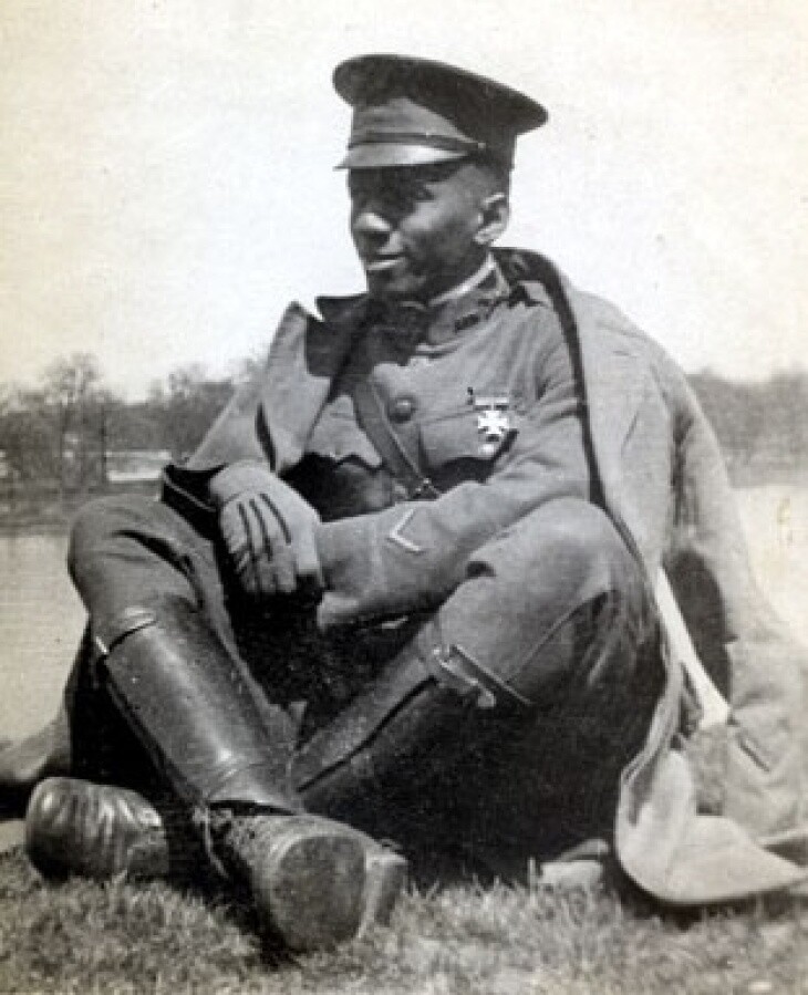 a Black man wearing a military uniform, knee-high leather boots, a military cap and a coat sits with his legs crossed on the ground
