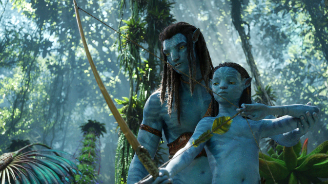 Sam Worthington and Jamie Flatters stand in a green, tropical forest with tall trees and lush plants. They both have blue skin, pointed ears, and long black hair, signature features of the Na'vi, the native inhabitants of the planet Pandora. Worthington stands behind the shorter and younger Flatters. Flatters holds a bow in his right hand with the strong of the bow and an arrow with leaf feathers in his left. His right arm is extended towards the ground and his left arm is bent as he draws back the arrow, preparing to fire. Worthington holds Flatters' left elbow up, guiding him, teaching him how to aim and shoot. They both look toward the ground where the arrow is aimed. 