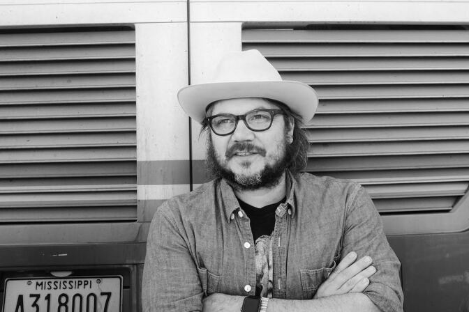 A  black and white image of Jeff Tweedy in a hat, glasses and button-down shirt leaning up against a truck with Mississippi plates.