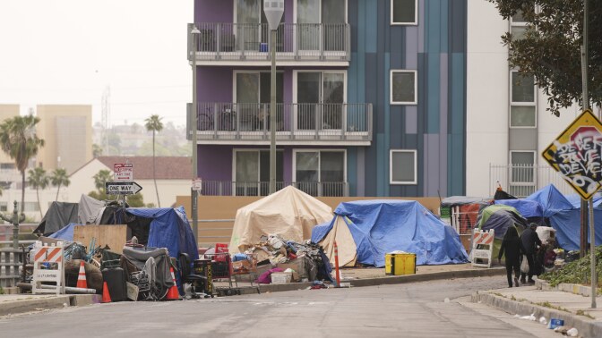 A group of tents along a sidewalk are next to an apartment building with balconies.