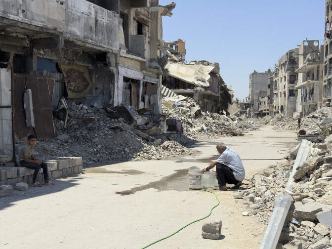 A man bends down, filling a jug with water from a pipe in the middle of a street. On all sides are buildings that are either completely or mostly destroyed.  